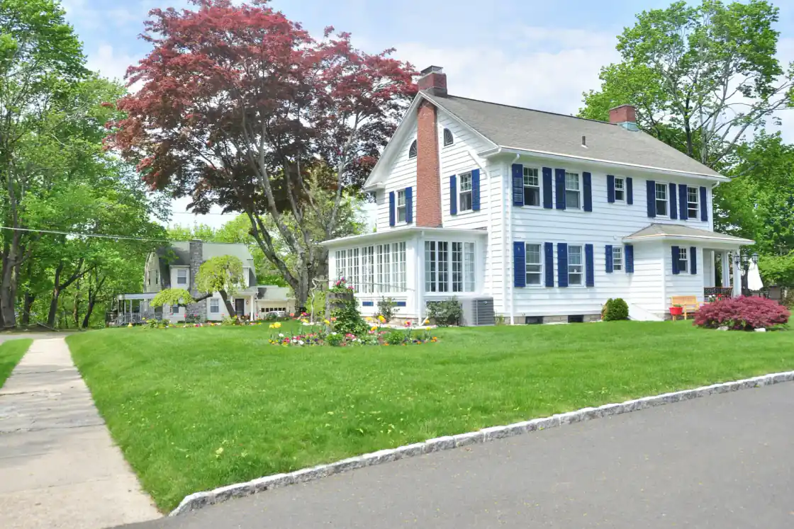 White colonial-style house with navy shutters, a brick chimney and a sunroom behind a green lawn in New England